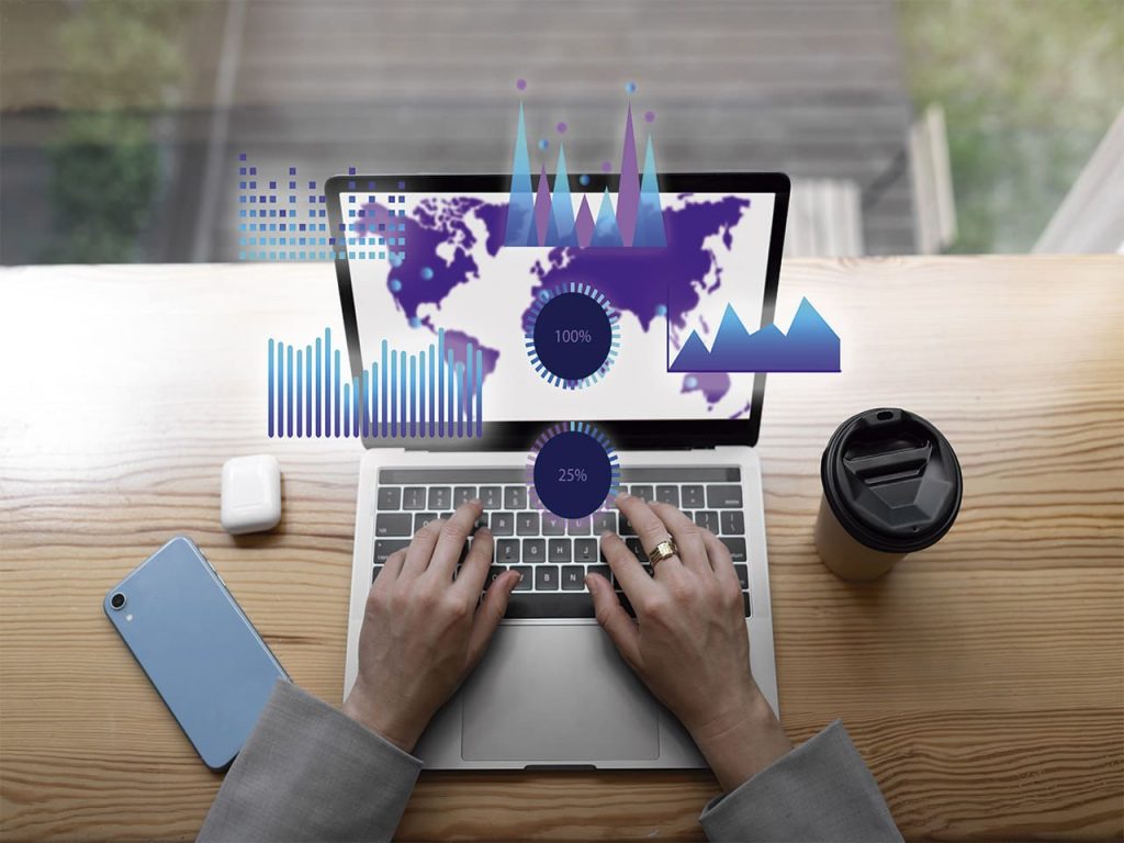 Imagem de uma pessoa trabalhando em um laptop com gráficos digitais sobrepostos na tela, incluindo mapas e estatísticas. A mesa de madeira tem um smartphone, fones de ouvido e um copo de café ao lado do computador, sugerindo um ambiente de trabalho focado em otimização de sites e otimização de sites.
